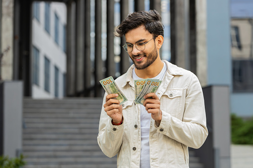 Happy excited Indian man counting money cash holding dollars banknotes bills budget count salary profit reward bribe outdoor. Successful rich Arabian Hindu guy in downtown city street. Business people
