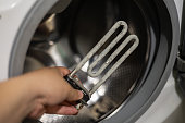 Close-up of a hand holding a heating element covered with limescale. Washing machine failure due to scale accumulation on the heating element. Washing machine heating element repair concept.