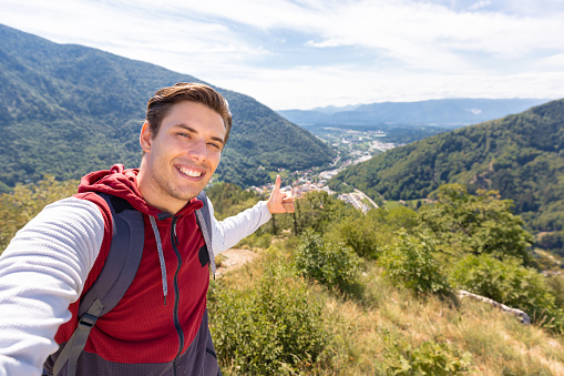 Smiling man taking a selfie video, talking and pointing at nature's beauty, mountain panorama in the background