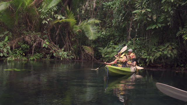 Senior adult Asian couple wearing casual clothing and life jacket having an adventure trip together in a mangrove forest and kayaking on a body of freshwater for recreation and relaxing under nature environment.