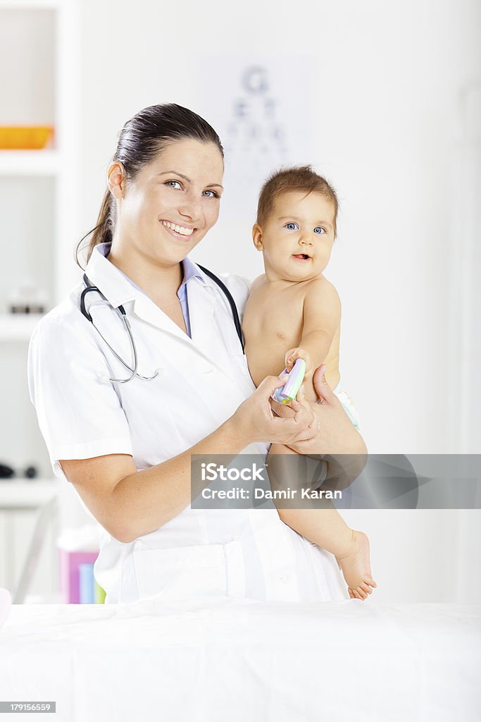 Female pediatrician with baby patient Female pediatrician standing in office. She is holding baby girl and smiling. Both of them are looking at camera Adult Stock Photo