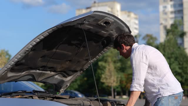Worried young man looking under the hood of breakdown car at outdoor in the city. Car repair. Transport and lifestyle concept. Real time