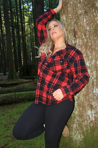 A Caucasian model in a forest leaning against a tree. She is wearing long blond hair, a red and black plaid shirt and black pants