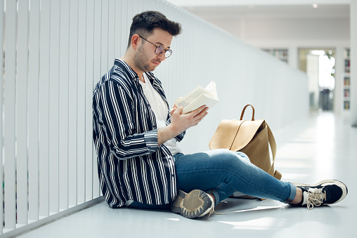 Man, student and reading with glasses and book for knowledge, education or learning at university. Male learner in library with books for thesis, project or assignment for scholarship at the campus