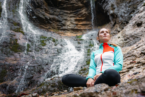 Young woman practicing yoga, sitting in om pose on the rock near the beautiful waterfall. Relax, nature, and wellbeing concepts.