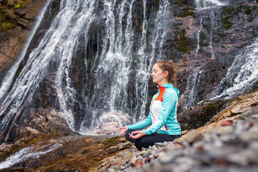 Fit young woman practicing yoga in beautiful waterfall scenery. Concepts of holistic experience, people and nature connection.