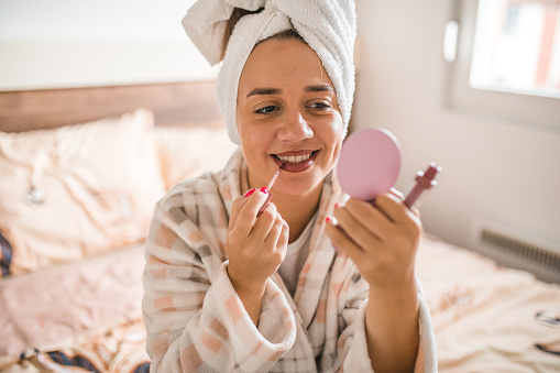 A smiling woman with a towel on her head is sitting on the bed in the bedroom and applying make-up