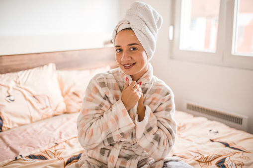 Portrait of smiling woman in bathrobe sitting on bed in bedroom and looking directly at camera