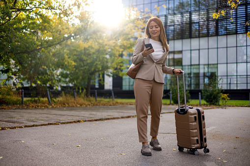 A mid-adult businesswoman is seen walking briskly with a suitcase while engaging with her mobile phone, a picture of efficiency and multitasking