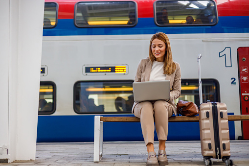 The photo shows a mid-adult businesswoman seated on a station bench, laptop open before her, epitomizing the remote work lifestyle