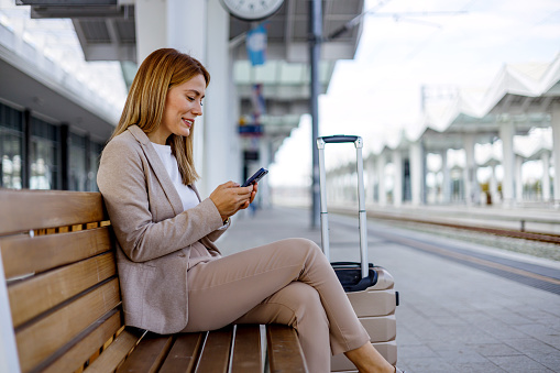 A mid-adult businesswoman is seen using her mobile phone while seated on a bench at the train station, effectively transforming a public space into a temporary office