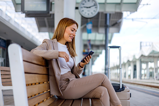 The image captures a mid-adult businesswoman seated on a train station bench, deeply engrossed in her phone, managing work or personal communications during her transit