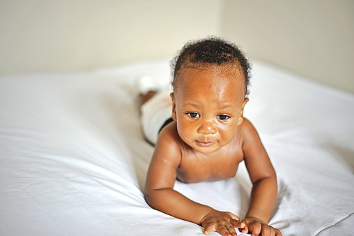 Childcare Concept. Portrait of cute little African  baby wearing bodysuit lying on white bedsheets at home. Black infant child crawling on bed in the bedroom.