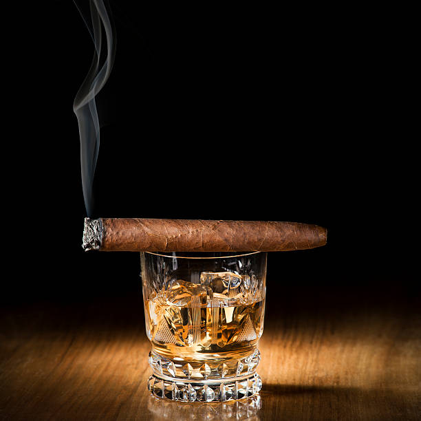 A burning cigar on top of a glass of alcoholic drink stock photo