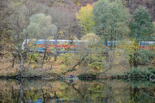 On the other side of the river, a fisherman is fishing. A blue train travels on a railway track. Beautiful views the autumn river Berounka, willow, forest and mountains, Hlasna Treban. Czech.