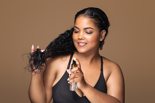 Haircare concept. Beautiful latin plus size lady applying hair spray for curls, standing over brown background and smiling. Body positive woman using new hairstyle product
