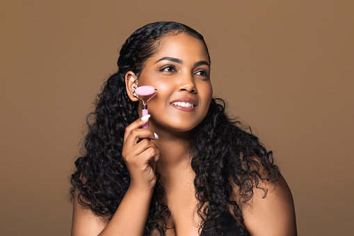 Skin lifting treatments. Beautiful brazilian plus size lady massaging face with jade roller, using beauty tool and smiling, standing isolated on brown background