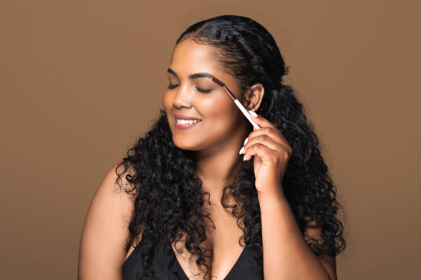 istock Charming latin body positive lady applying eyebrow mascara, making makeup, standing over brown studio background 1791762313