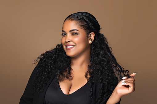 Happy brazilian chubby woman playing ideal curls after salon spa procedure, touching clean curly hair after hygienic routine against brown background, looking and smiling ar camera