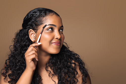 Beautiful brazilian body positive woman brushing eyebrows holding brush and looking aside at free space, enjoying beauty routine, brown studio background