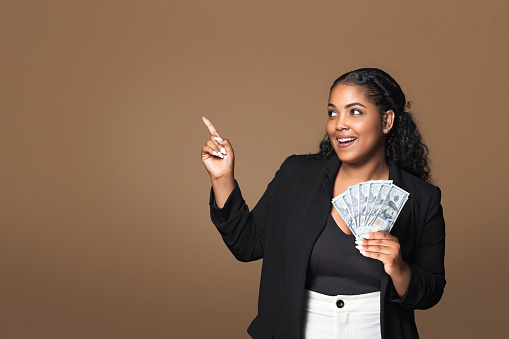 Happy latin plus size lady holding dollar cash fan in her hand and pointing aside at free space, celebrating financial success and profit, standing over brown studio background