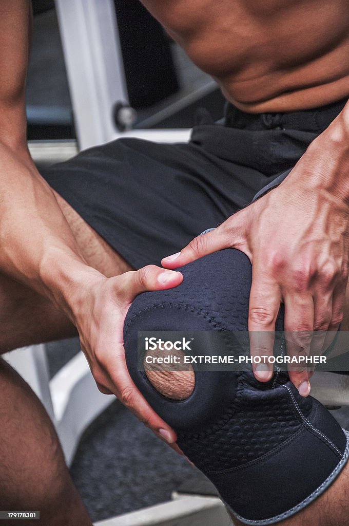 Knee pain Unknown male athlete holding injured knee with sports bandage while sitting on the exercise machine in the gym. Activity Stock Photo