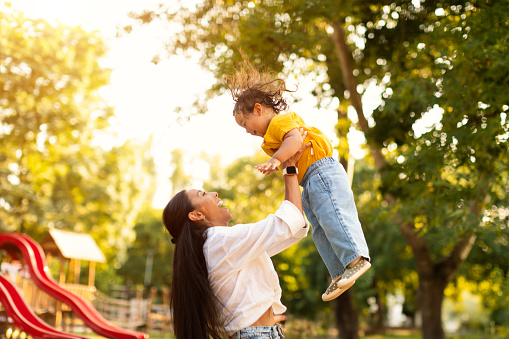 Joyful Park Playtime With Baby. Asian Mommy Entertaining And Delighting Her Little Toddler Daughter, Lifting Her Playfully into the Air Amidst Outdoor Playground. Happy Family, Motherhood Moments