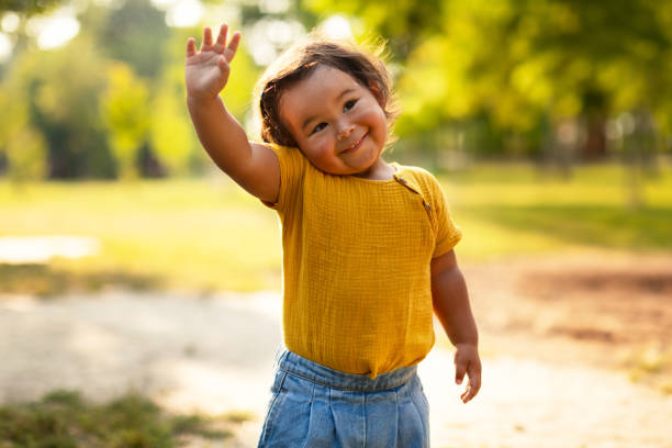 Adorable Little Baby Girl In Casual Waving Hand Standing Outdoor Hello. Adorable Little Baby Girl In Casual Waving Hand And Smiling To Camera Standing Outdoor, Enjoying Joyful Summer Adventures Amidst Beauty of Nature, Portrait Of Toddler Gesturing Hi Greeting kid waves stock pictures, royalty-free photos & images