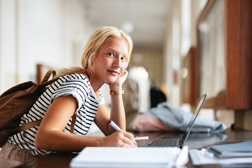 A portrait of a smiling young Caucasian woman writing something down while being at university.