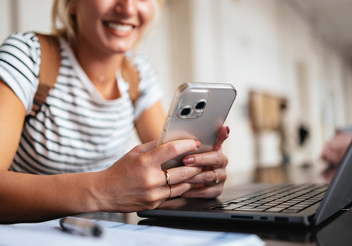 A close up view of a smiling young Caucasian woman using her smartphone while being at university. (defocused)