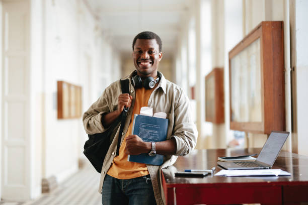 A Happy Handsome Student Holding A Book And Looking At Camera While Standing In The College Corridor