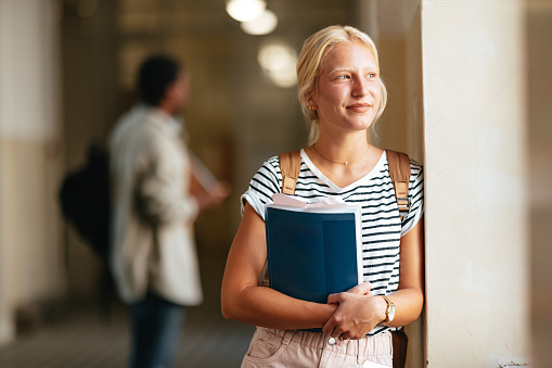 A smiling Caucasian female holding a book while standing at school and contemplating.