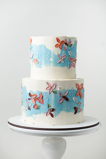 Birthday two tier cake with white chocolate frosting decorated with blue smears and multicolored pastel cream flowers on the white background.