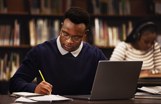 Student, man and writing in library for studying, research and planning in university, scholarship and education. African person with online test and computer for language, e learning or philosophy