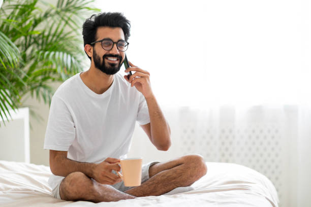 jovem indiano sentado na cama, falando no celular e bebendo café - hotel domestic room telephone technology - fotografias e filmes do acervo