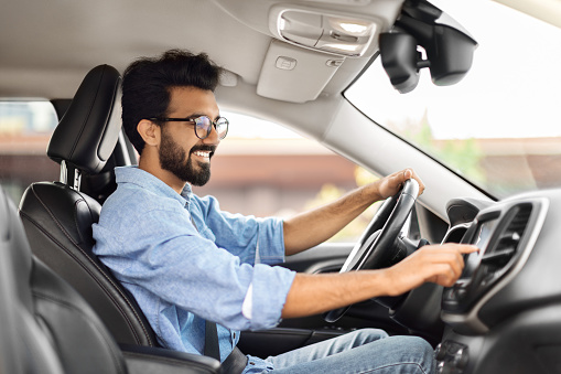 Road playlist. Side view of happy young indian man driving car and listening to music radio on auto audio system, checking automobile options during test drive, pushing button on dashboard