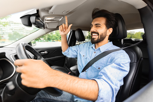 Emotional millennial handsome bearded arab guy driver having fun during car trip, listening to music and cherfully singing, moving his hand, shot from open side window of auto
