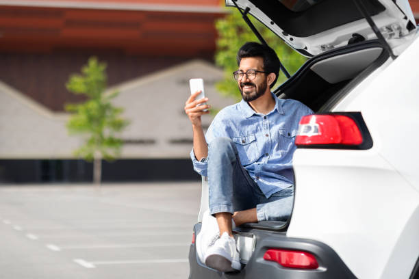 istock Cheerful indian guy sitting in open car trunk, using smartphone 1791837720