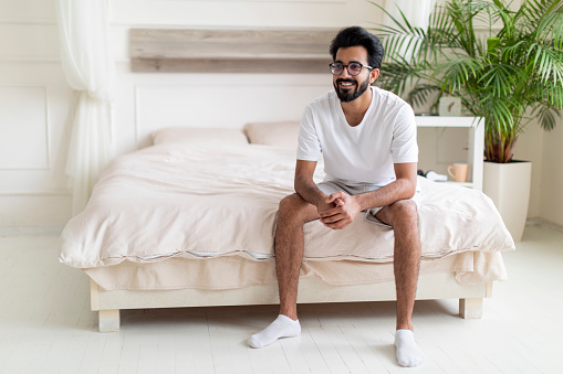 Portrait Of Happy Young Indian Man Sitting On Bed In Bedroom After Waking Up, Cheerful Smiling Eastern Guy Resting In Pajamas And Looking Away, Millennial Male Enjoying New Day, Copy Space