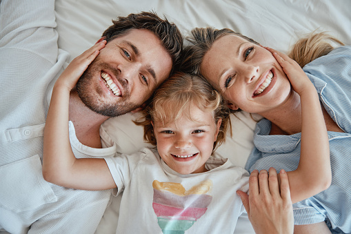 Family, children and face with a girl, mother and father lying on a bed together in their home in the morning. Portrait, bedroom and bonding with a man, woman and daughter in the bedroom from above