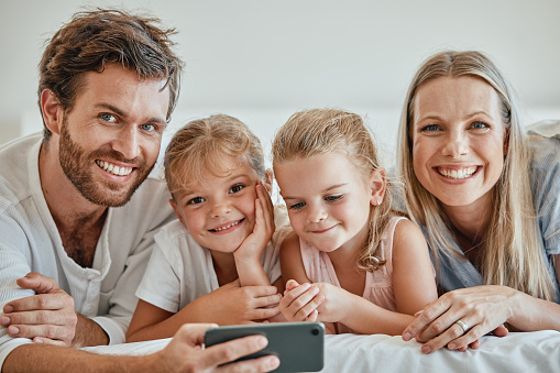 Family, phone and bed with a girl, sister and parents bonding together in the bedroom of the home in the morning. Children, portrait and mobile with a man, woman and daughter siblings lying on a bed