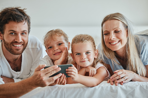 Happy, family and relax with phone on the bed in fun entertainment streaming together at home. Mother, father and children relaxing in the bedroom with smile for online shows on smartphone indoors