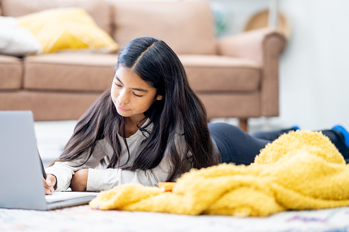 A young adolescent of Hispanic decent, lays on her living room floor with her laptop open in front of her as she attends school virtually.  She is dressed casually and taking notes.