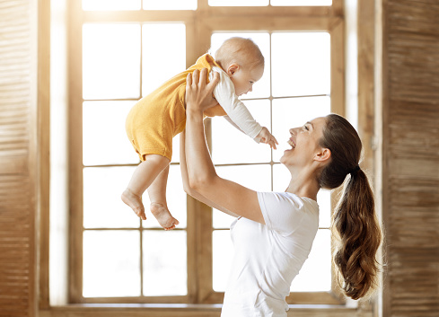 Motherhood, parentthood, child care concept. Loving happy young mother lifting up her adorable cute little baby, mom and kid bonding at home, standing by window. Love, affection, family