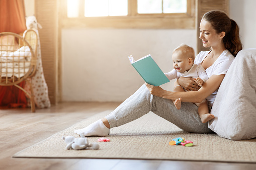 Happy loving young mother and her cute baby son reading book together at home while relaxing on floor in kids room, free space, sun flare. Kids education, child care, motherhood concept