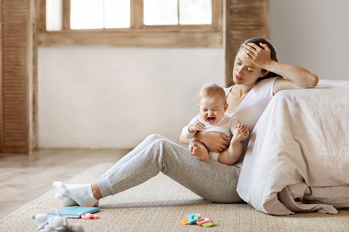 Tired infant baby cries in mother hands, depressed unhappy exhausted mom sitting on floor with crying little child on her lap, bedroom interior, copy space. Postnatal postpartum depression concept