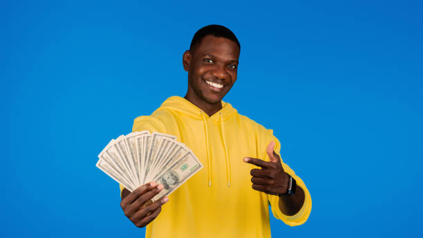 istock Cheerful young black guy points finger at many money dollars, isolated on blue background 1791909816