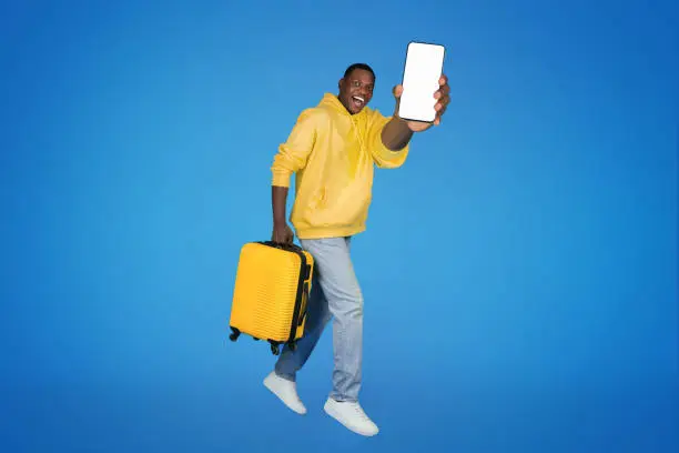 Photo of Glad young african american guy goes with suitcase, freezes in air, shows smartphone with empty screen