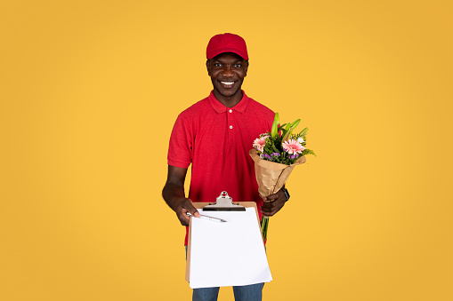 Cheerful millennial african american guy courier hold bouquet of flowers, gives clipboard for sign, isolated on orange studio background. Delivery of gift at home, work and service for holiday
