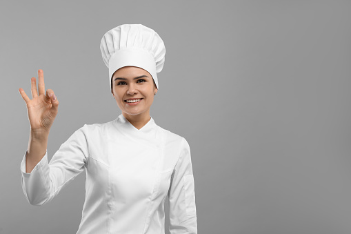 Happy female chef wearing uniform and cap showing ok gesture on light grey background. Space for text
